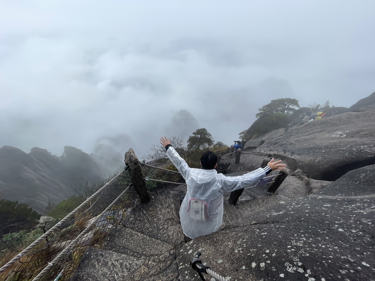 雨の中、霧の雲海を前に天都峰の岩場で両手を広げる登山者