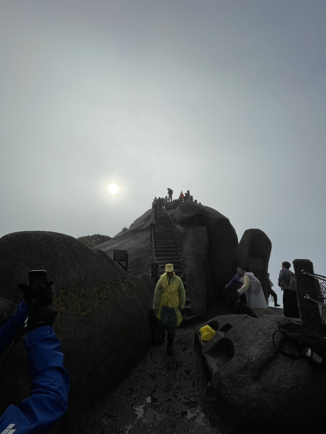 霧の向こうにうっすらと太陽が見える天都峰の山頂と、記念撮影をする登山者たち