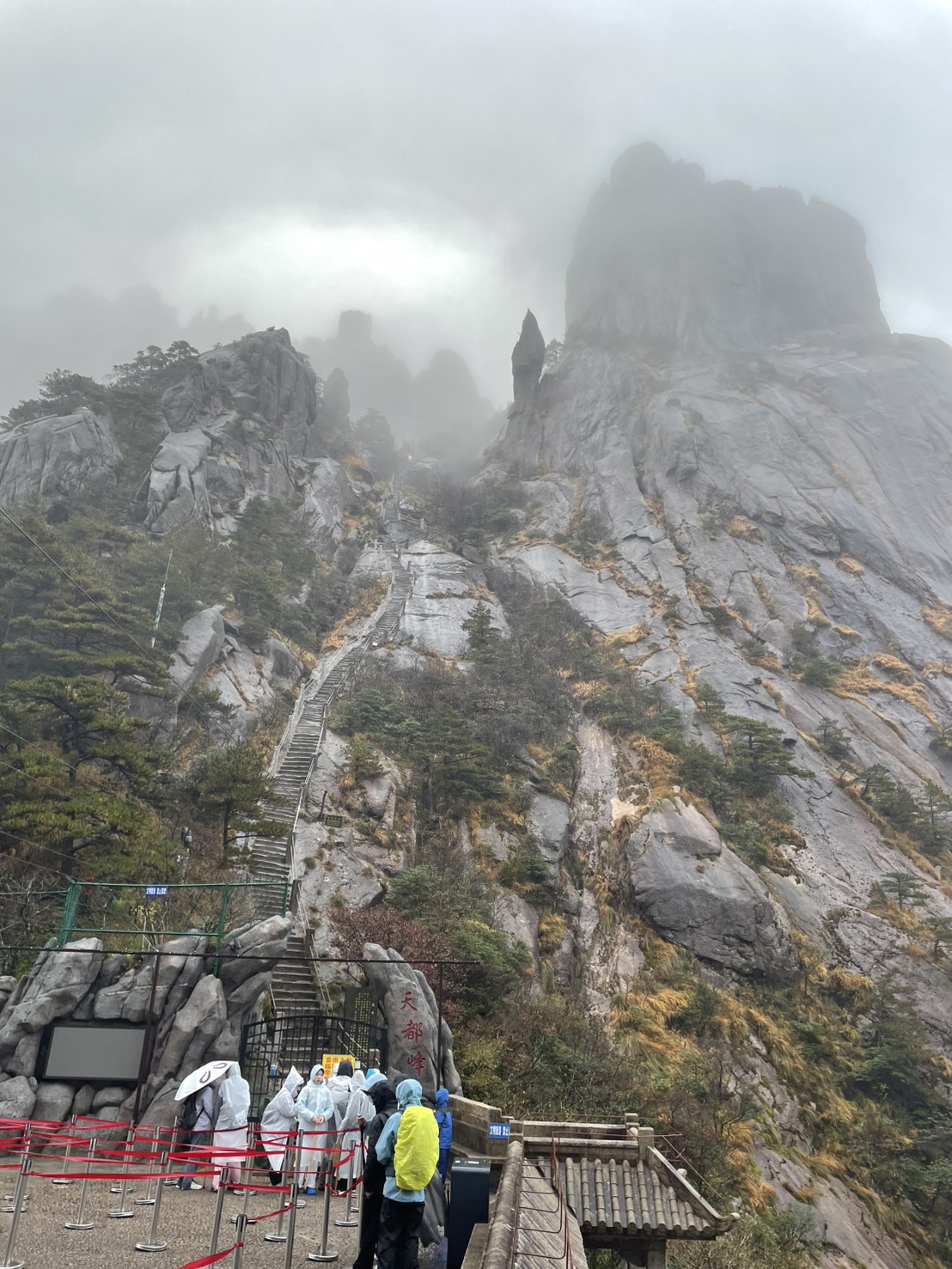 雨の中、カッパ姿の人々が並ぶ天都峰の登山口と、岩山に続く長い石段
